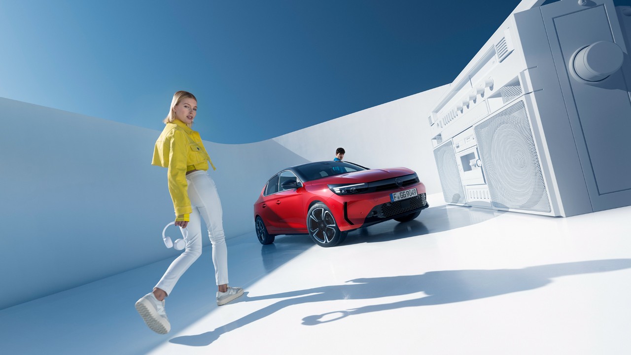 Vrouw naast een Nieuwe Opel Corsa in rood met een zwart dak en een man die instapt aan de bestuurderzijde