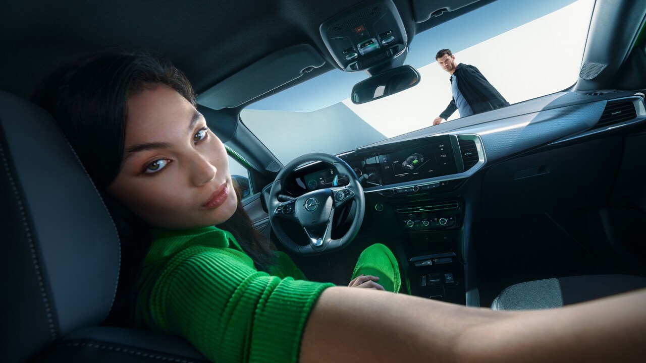 Een vrouw in de bestuurdersstoel van een Opel Mokka Electric kijkt achterom terwijl er buiten een man loopt